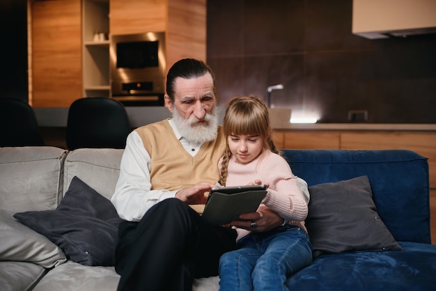 Nonno con sua nipote che per mezzo del pc della compressa a casa accogliente. la bambina insegna a suo nonno a usare dispositivi intelligenti.