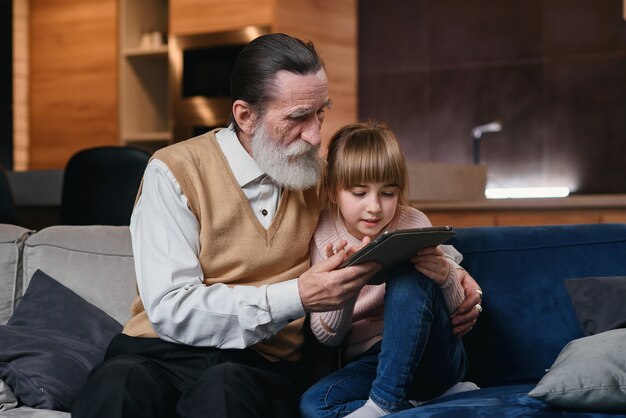 Nonno con sua nipote che per mezzo del pc della compressa a casa accogliente. la bambina insegna a suo nonno a usare dispositivi intelligenti.