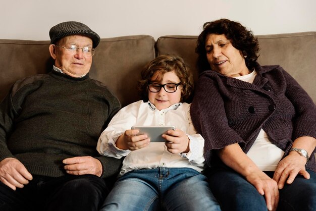 Photo grandparents looking at grandson using smart phone while sitting on sofa at home