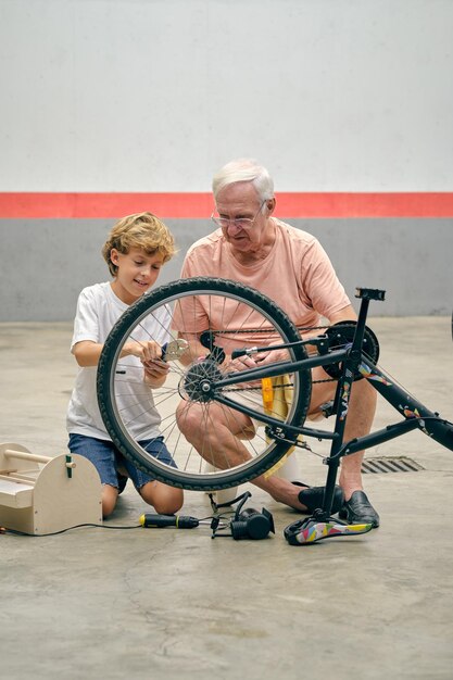 Grandson in sport outfit sitting on concrete floor near bicycle and repairing wheel under grandfather control