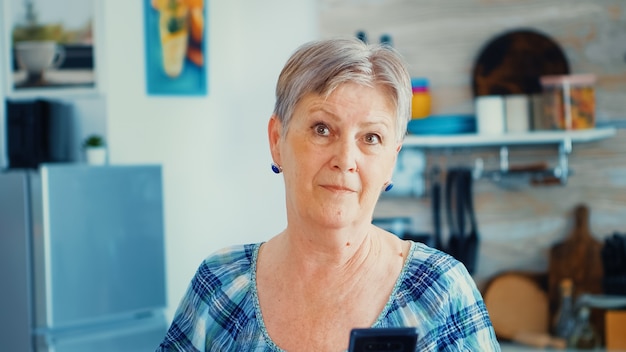写真 朝食時にキッチンでスマートフォンを使用しながらカメラを見ているおばあちゃん。現代のスマートフォンインターネット技術を使用した本物の高齢者。世界とつながるオンラインコミュニケーション、シニアl