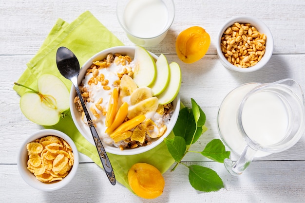 Гранола и вегетарианский йогурт с кусочками яблока, абрикоса, банана и кувшин молока на белом фоне деревянные.