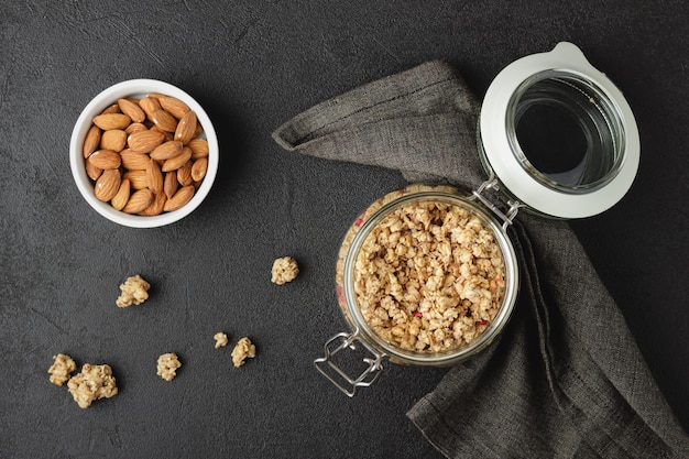 Granola in jar, almonds in white bowl and dark napkin. Healthy breakfast on black table. Top view.