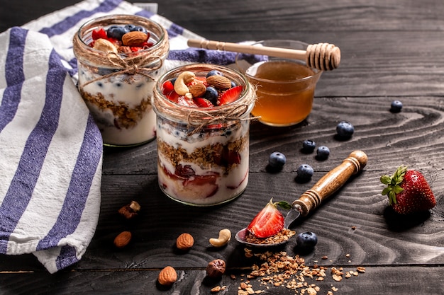 Granola Parfait met yoghurt, haver granola, verse bessen, honing in hoge glazen pot, tekst recept op houten achtergrond.