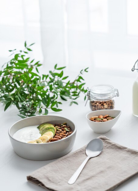 Granola with banana and kiwi on a white table with a green branch against the background of a window