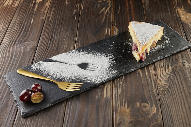 Grape cake with powdered sugar and grapes on a black stone board and wooden background. French gourmet food