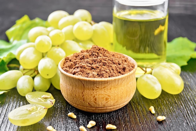 Grape seed flour in a bowl, oil in a jar and grapes berries on dark wooden board background