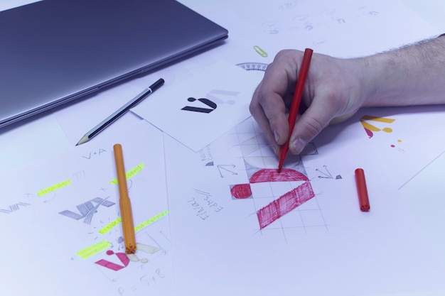 Photo graphic designer designs a logo against a background of sketches and drawings on a table. printed logos on paper in a studio with a laptop.