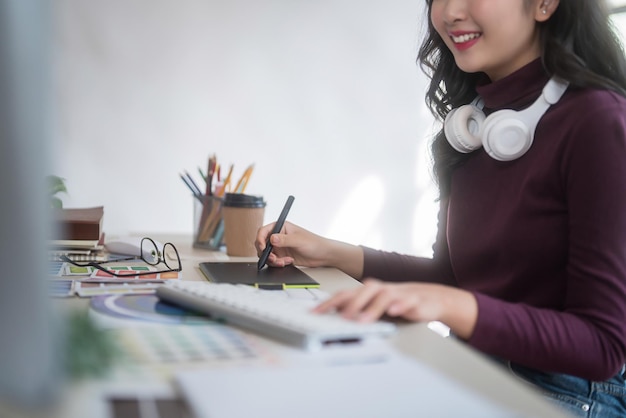 写真 グラフィックデザイナーのフリーランス女性はヘッドフォンを首に付けデジタルタブレットでブランドのロゴをスケッチしホームスタジオで技術機器でグラフィックデザインをしている間コンピュータでタイピングします