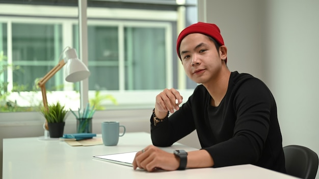 Graphic designer in red wool hat working on digital tablet and looking at camera while sitting at his workplace.