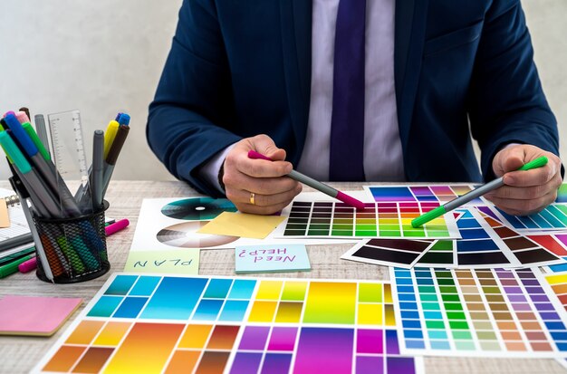 Graphic or interior young designer in suit choosing color from swatch sample or catalogue palette guide at workplace. graphic designer with paint color palette samples at table, close up