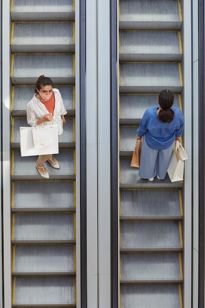 Graphic top down view at two women riding escalator in opposite directions while shopping in mall, focus on woman wearing mask looking up at camera, copy space