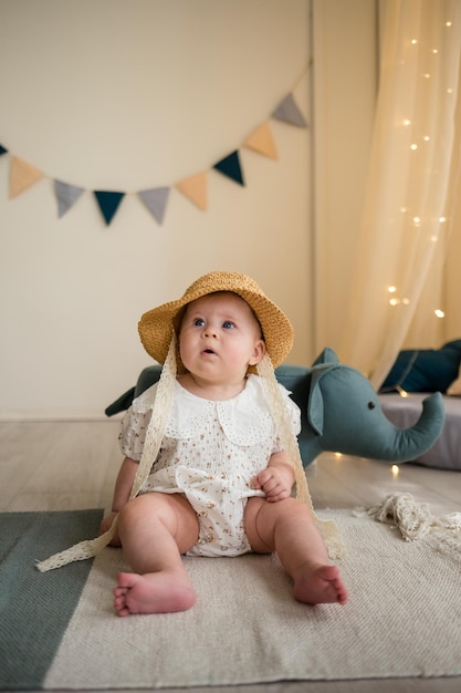 Grappig babymeisje in een bodysuit en een strohoed zit op de grond in de kinderkamer