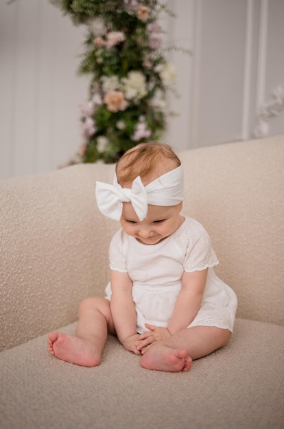 Grappig babymeisje in een witte jurk met een hoofdband zit in een beige fauteuil in een witte kamer