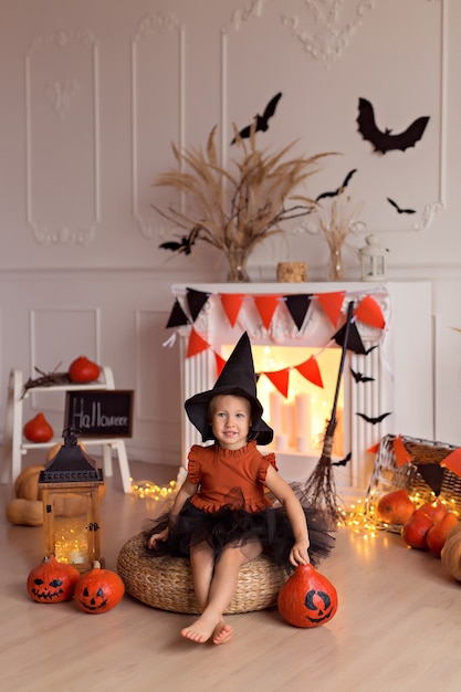 Foto grappig babymeisje in halloween-heksenkostuum met pompoenhefboom en bezem binnenshuis