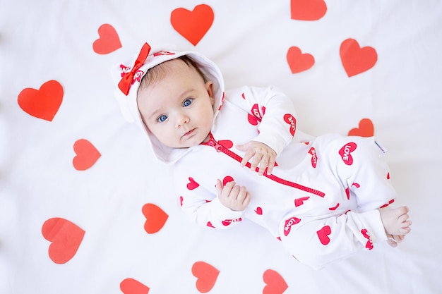 Grappig babymeisje met rode harten op een bed op een wit katoenen bed dat valentijnsdagconcept glimlacht of lacht