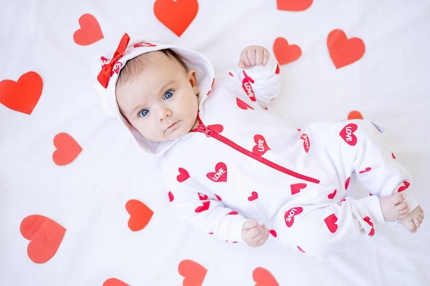 Grappig babymeisje met rode harten op een bed op een wit katoenen bed dat valentijnsdagconcept glimlacht of lacht