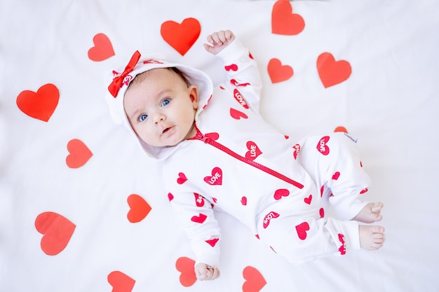 Grappig babymeisje met rode harten op een bed op een wit katoenen bed dat valentijnsdagconcept glimlacht of lacht