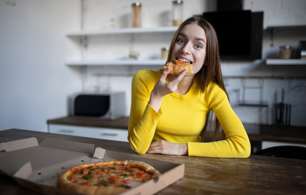 Grappig donkerbruin meisje dat in gele sweater pizza eet bij keuken