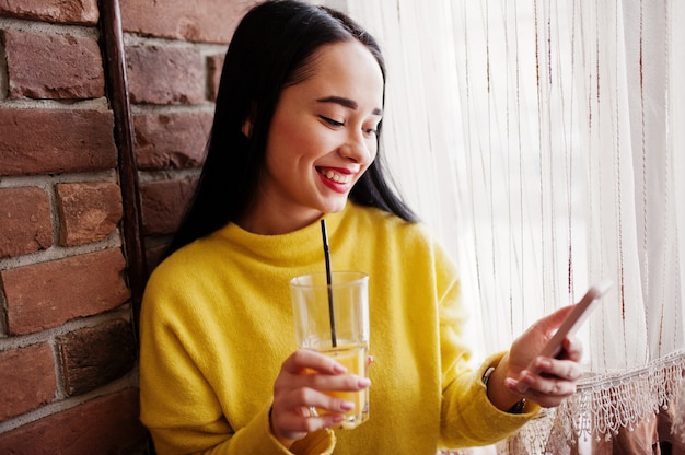 Foto grappig donkerbruin meisje in geel sweater het drinken sap en het bekijken mobiele telefoon restaurant.