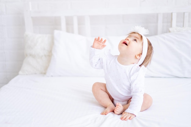 Grappig gelukkig babymeisje zittend op het bed in witte kleren en haar handen opstekend een klein kind opzoekend op een katoenen bed thuis glimlachend het concept van kinderartikelen