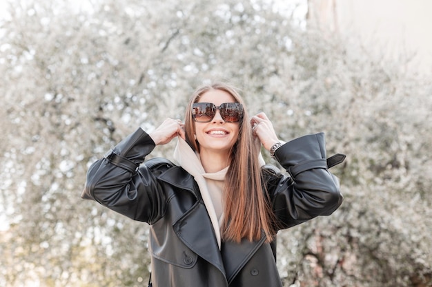 Grappig gelukkig mooi modieus meisjesmodel met een glimlach met zonnebril in een modieuze zwarte jas met een hoodie, draag een capuchon, geniet van en wandel in een bloeiend park