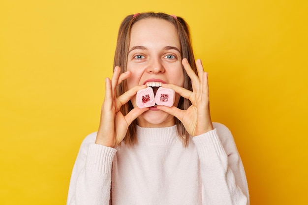 Foto grappig gelukkig vrolijk jong meisje met paardenstaarten in trui bijten marshmallow poseren geïsoleerd over gele achtergrond kijken naar camera desserts eten