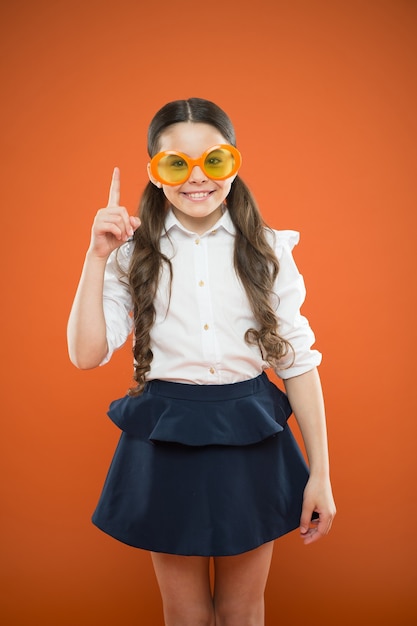 Grappig idee. Klein meisje in grappige bril wijzende vinger omhoog op oranje achtergrond. Grappig kind dat een zonnebril met kleurenfilter draagt. Mijn ogen worden grappig.