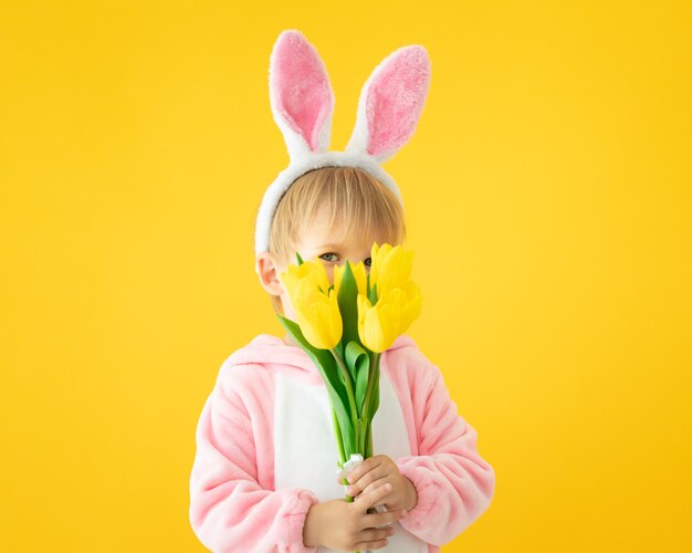 Grappig jong geitje dat paashaasoren draagt en een boeket tulpen tegen gele muur houdt.