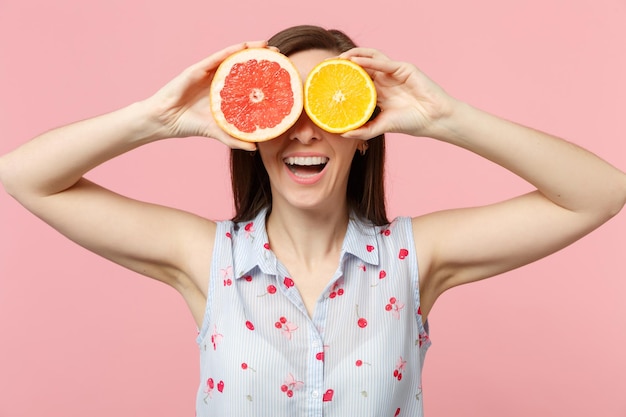 Grappig jong meisje in zomerkleren die ogen bedekken met helften van vers rijp grapefruit oranje fruit geïsoleerd op roze pastel achtergrond. Mensen levendige levensstijl ontspannen vakantie concept. Bespotten kopie ruimte.