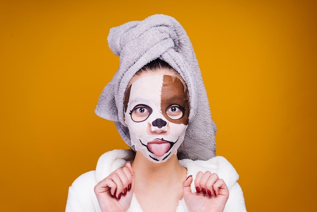 Grappig jong meisje met een handdoek op haar hoofd toont haar tong op haar gezicht een handig hydraterend masker met een hondengezicht