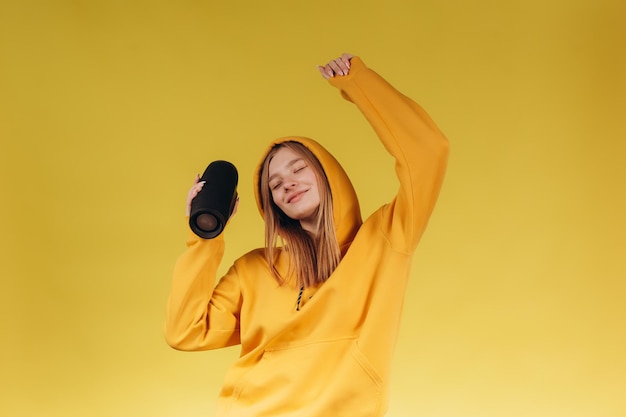 Grappig jong meisje op zoek zwaaiende hand met draagbare draadloze bluetooth-muziekluidspreker geïsoleerd op gele achtergrond Meisje in een gele hoodie