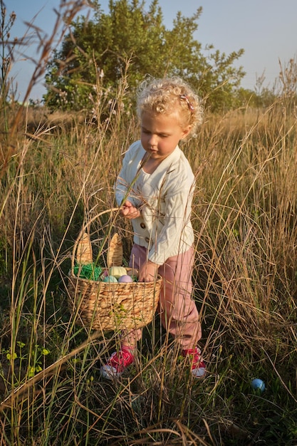 Grappig klein meisje in witte trui die geverfde paaseieren verzamelt in rieten mand op met gras begroeide groene weide in de zonnige lente natuur