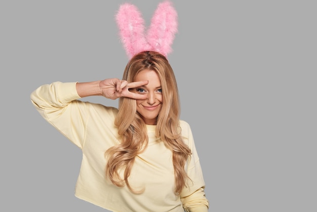Foto grappig konijn. aantrekkelijke jonge glimlachende vrouw in roze konijnenoren die gebaren en naar de camera kijken terwijl ze tegen een grijze achtergrond staat