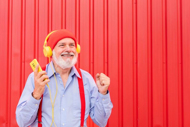 Grappig lachende gepensioneerde man met bretels luisteren naar muziek met koptelefoon dansen geïsoleerd op rode achtergrond