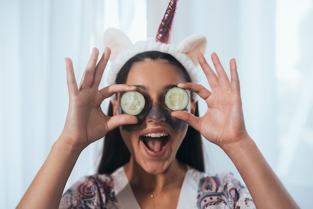 Grappig lachende spa-vrouw met vers gezichtsmasker houdt komkommers vast