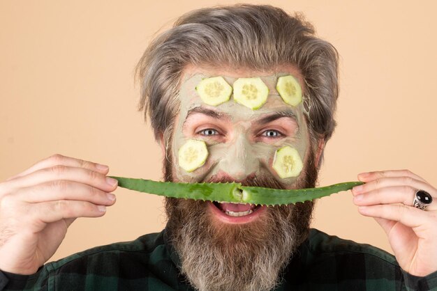 Grappig mannelijk model met kleimasker en plakjes komkommer, aloë vera snor op gezicht. Spa, dermatologie, wellness en gezichtsbehandeling concept. Man met cosmetisch hydraterend masker, geïsoleerd.