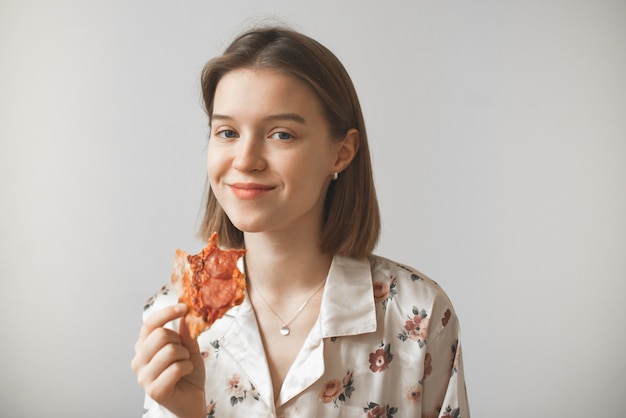 Grappig meisje in een pyjama met een stuk pizza in haar handen kijkt naar de camera en glimlacht met een grijns.