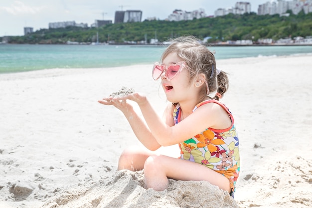 Grappig meisje poseren in roze zonnebril met zand in haar handen. Kinderspellen op het strand.