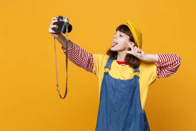 Grappig meisje tiener in franse baret doen selfie geschoten op retro vintage fotocamera, tong, overwinningsteken geïsoleerd op gele achtergrond tonen. mensen emoties, lifestyle concept. bespotten kopie ruimte.