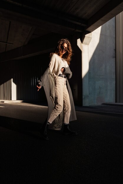 Grappig mooi meisjesmodel met krullend haar in modieuze kleding met een vintage lange jas die op straat in het zonlicht loopt. Stedelijke vrouwelijke stijl en elegante mode