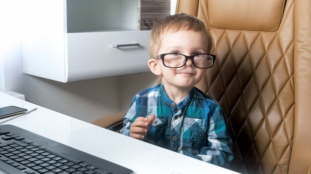 Grappig portret van een lachende peuterjongen in een bril die achter een bureau op kantoor zit.