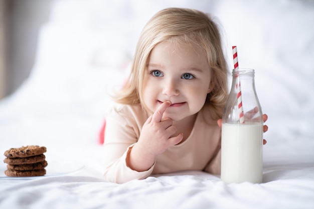 Grappig schattig klein kind meisje 3-4 jaar oud consumptiemelk in glazen fles en chocolade koekjes eten in stapel liggend in bed op witte deken close-up. Ontbijt tijd. Goedemorgen. Jeugd.