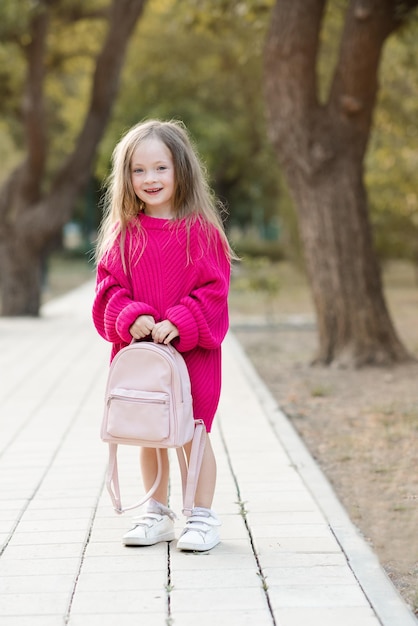 Grappig schattig meisje van 5-6 jaar oud draagt een roze gebreide trui met een rugzak die op de weg in het park staat