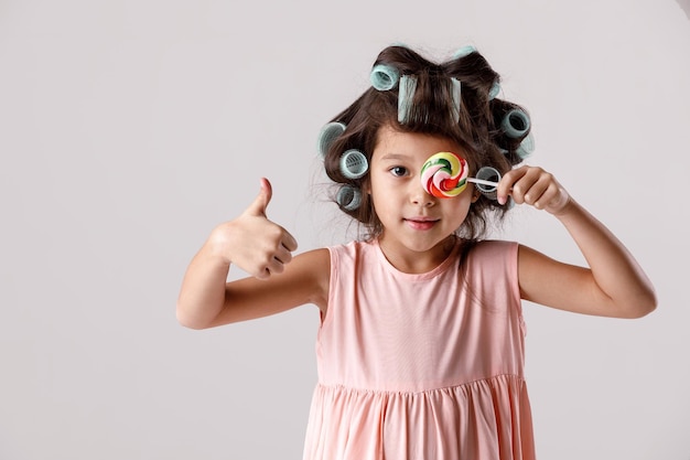 Grappig tevreden klein kindmeisje in roze jurk en haarkrulspelden die lolly op grijze achtergrond houden.