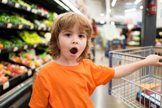 Grappig winkelend vrolijk mooi kindjongen in supermarkt koopt groenten, gezond voedsel voor kinderen