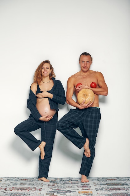 Grappig zwanger meisje met haar man speelt Zwangere vrouw houdt haar buik vast en haar man houdt een pompoen vast in plaats van zijn buik en tomaten op zijn borst