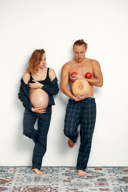 Grappig zwanger meisje met haar man speelt Zwangere vrouw houdt haar buik vast en haar man houdt een pompoen vast in plaats van zijn buik en tomaten op zijn borst