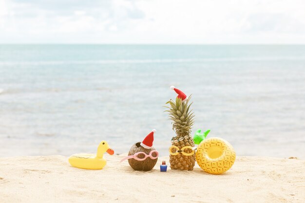 Grappige aantrekkelijke ananas en kokos in stijlvolle zonnebril op het zand tegen turkooizen zee. kerstmutsen dragen.