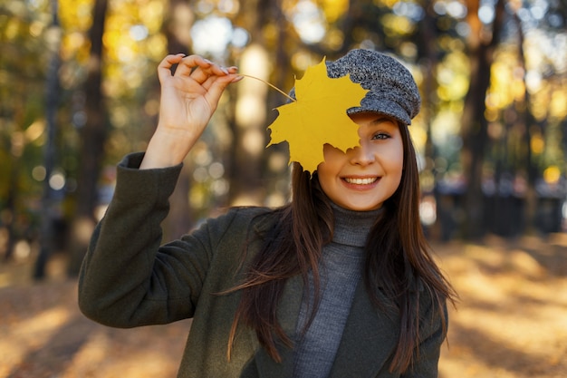 Grappige aantrekkelijke mooie jonge gelukkig vrouw met een glimlach in een vintage hoed en jas heeft betrekking op haar gezicht met een gele Herfstblad en geniet van een zonnige dag